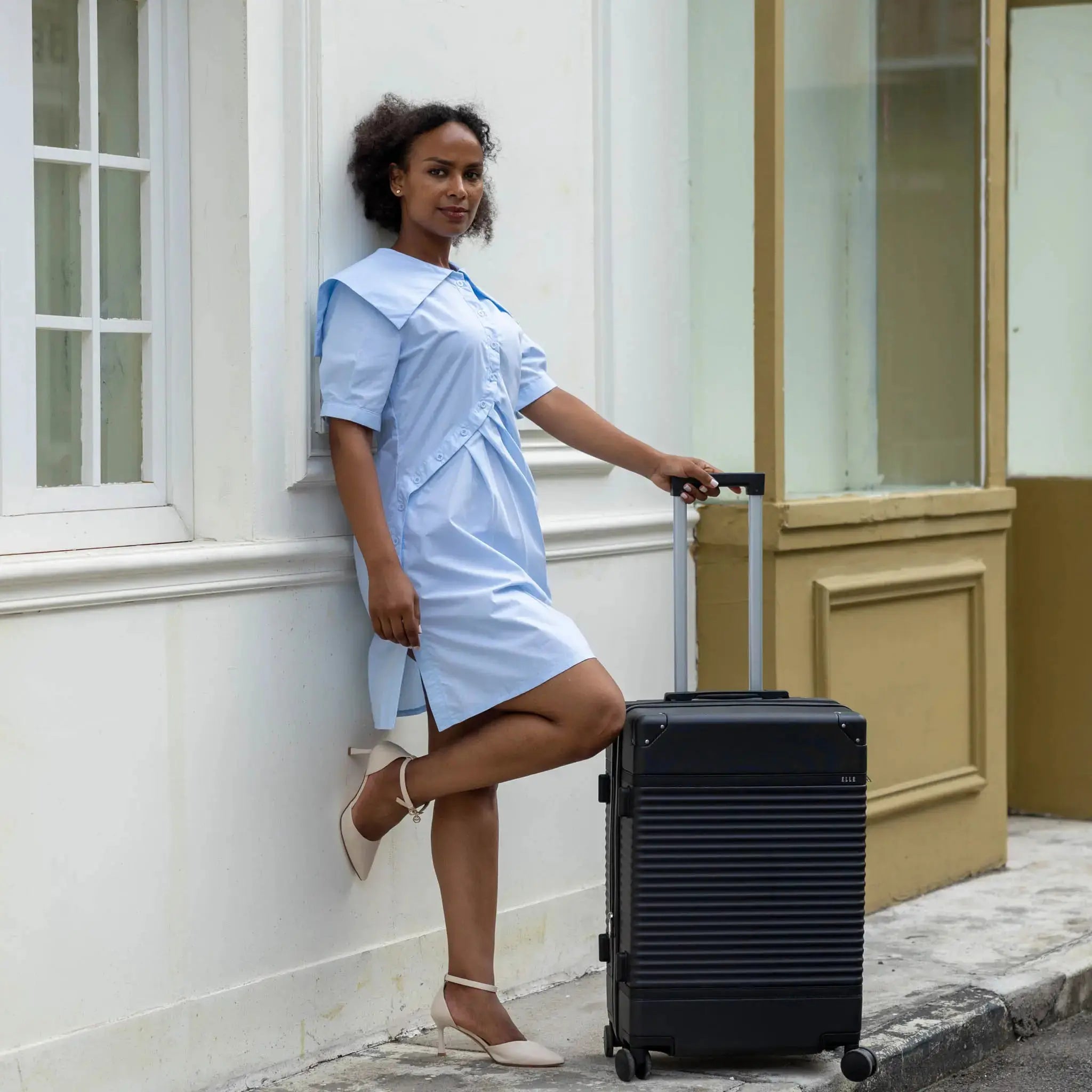 Woman in a light blue dress standing next to a black suitcase on a street.