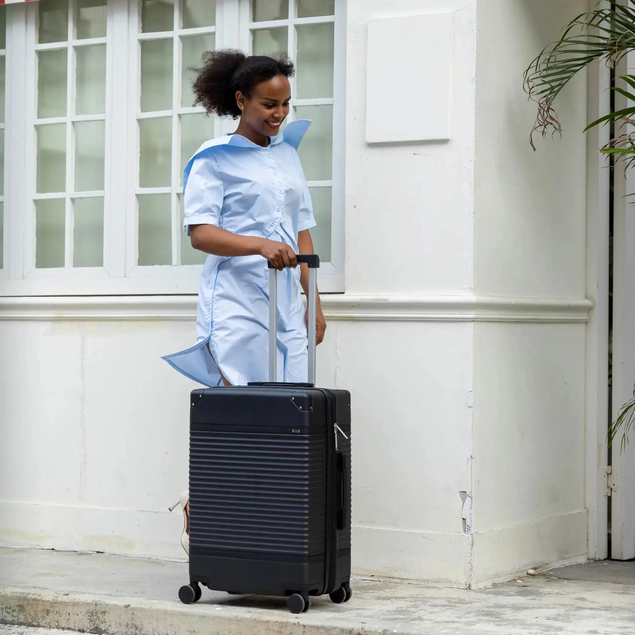 Woman in a light blue outfit pulling a black suitcase outside a building.