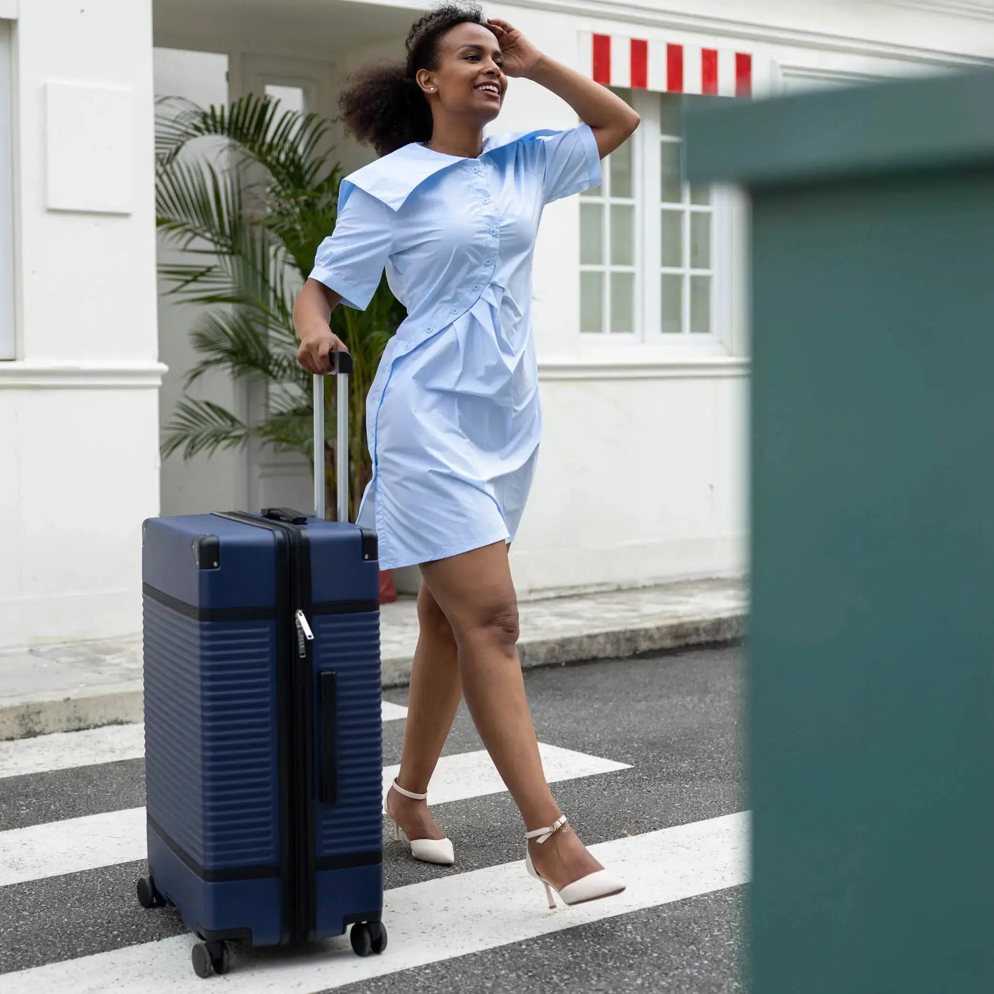 Woman in a light blue dress pulling a blue suitcase on a street.