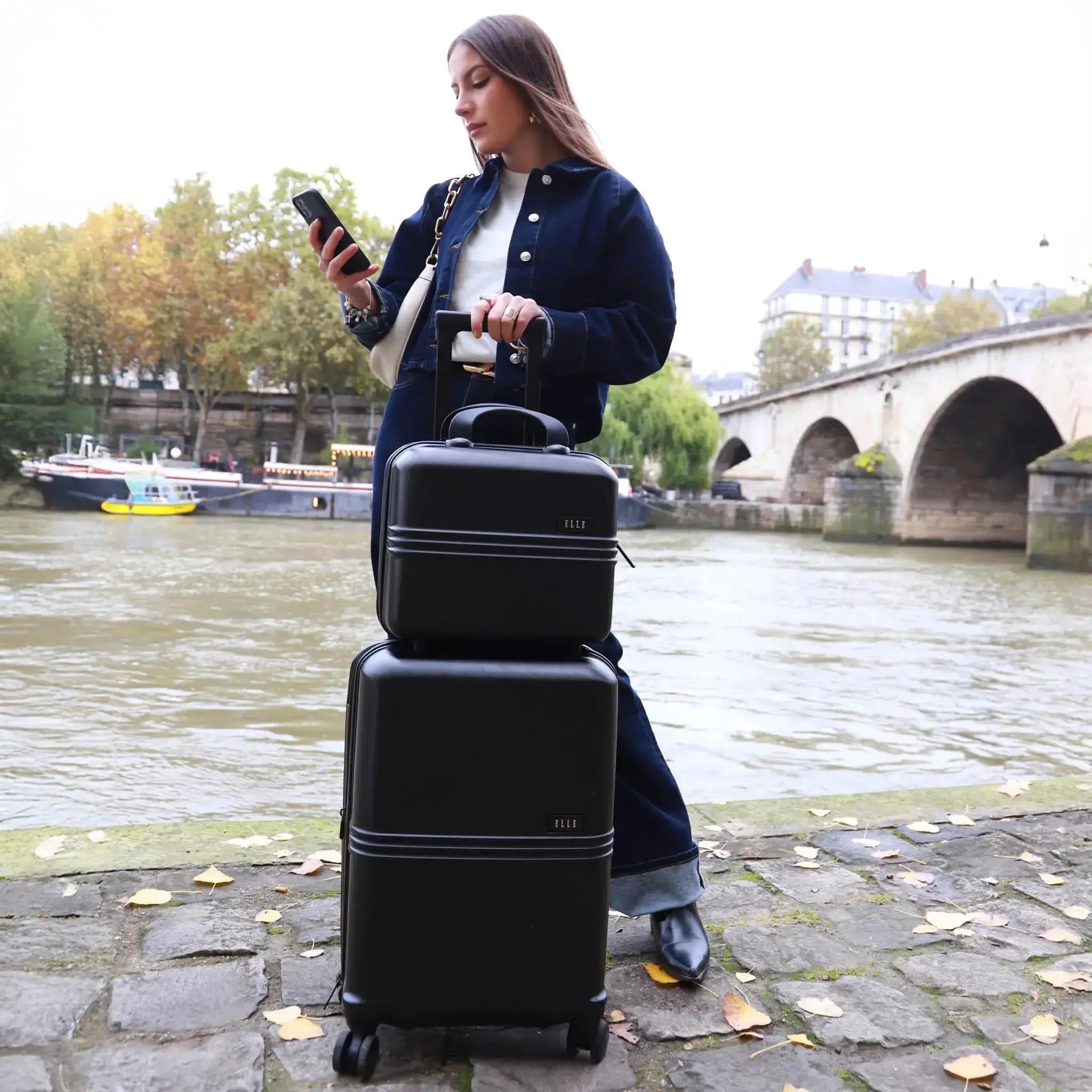 Woman with two black suitcases by a river with a bridge in the background