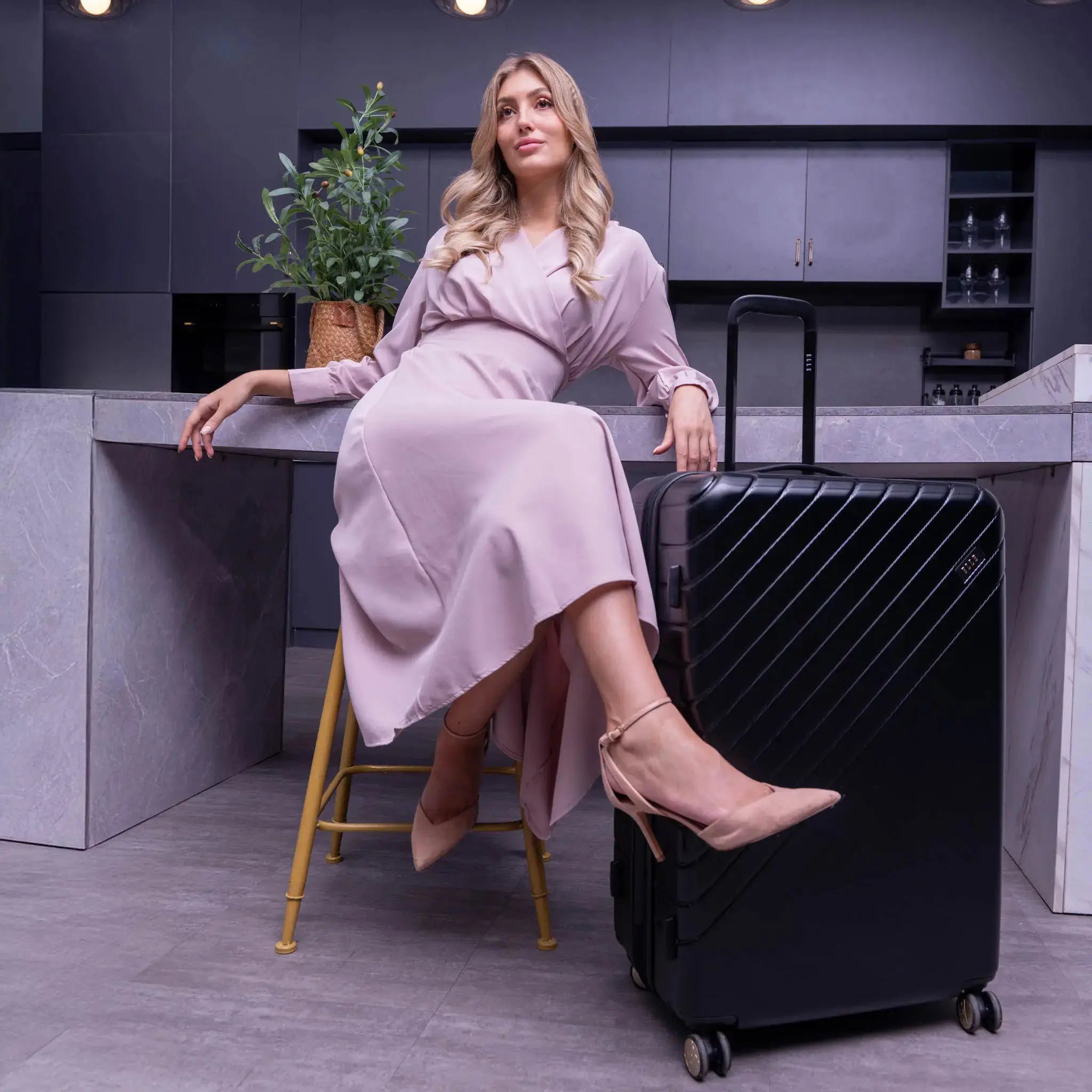 Woman in a pink dress sitting on a black suitcase in a modern kitchen.