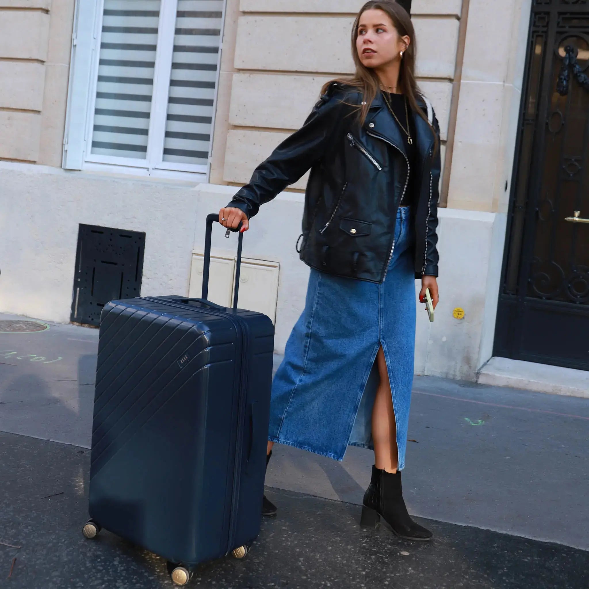 Woman in black leather jacket and blue denim skirt pulling a dark suitcase on a street.