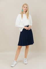 Woman wearing a white sweater and navy pleated skirt on a beige background