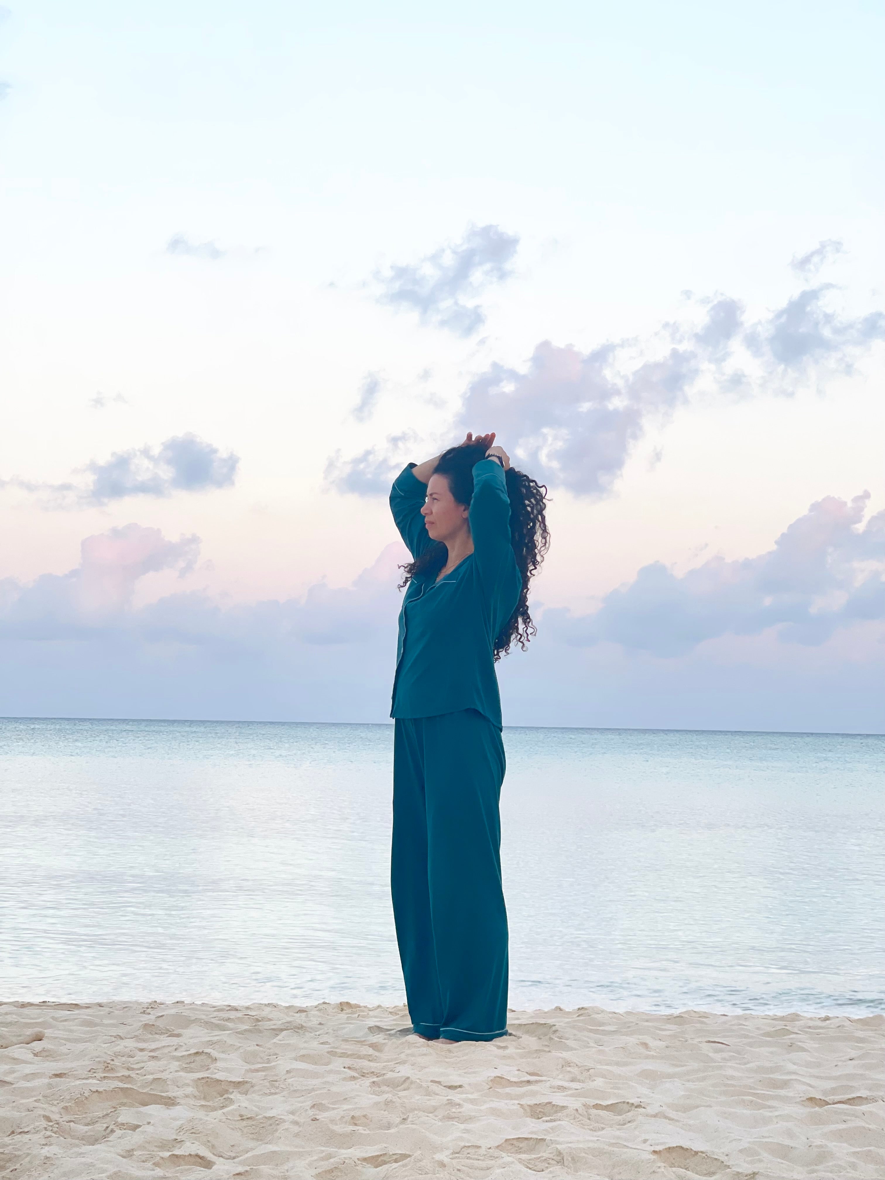 Person in teal outfit standing on a beach with ocean and cloudy sky in the background