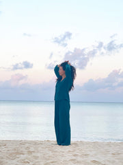 Person in teal outfit standing on a beach with ocean and cloudy sky in the background
