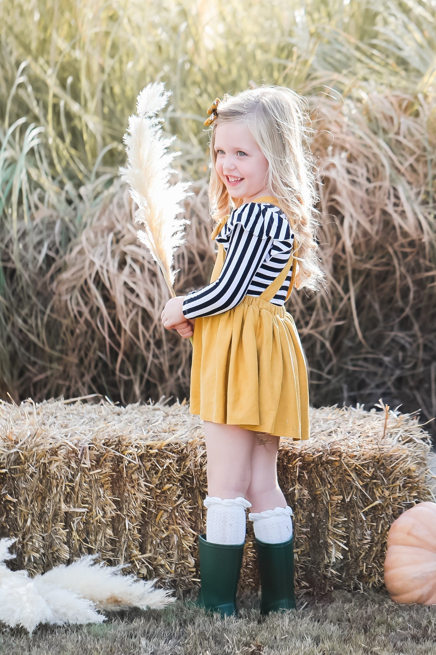 Young girl in a yellow dress and striped sweater standing next to pumpkins and hay bales.