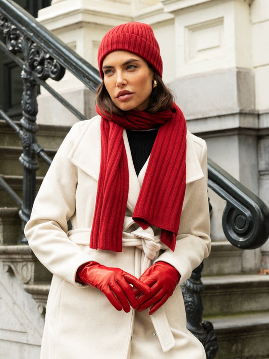 Woman wearing a red beanie, scarf, and gloves with a white coat against a stone building background.