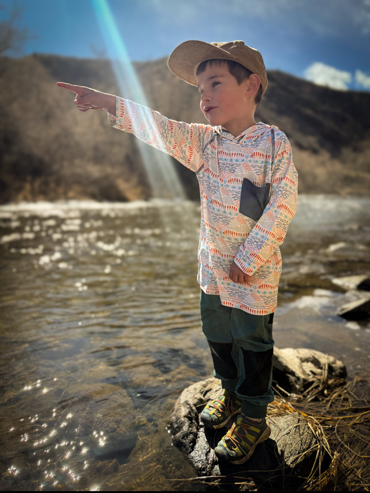 Child standing by a river, pointing towards the water with mountains in the background
