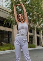 Woman in light gray tank top and pants stretching outdoors with trees and building in the background