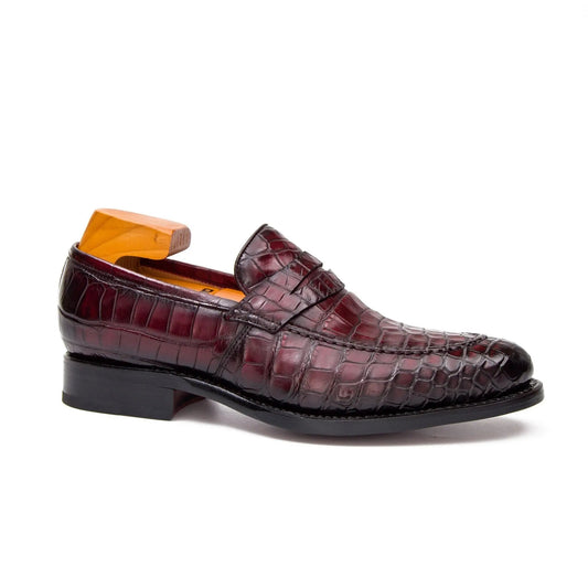 A single men's loafer in glossy embossed crocodile leather, shown in a side profile on a white background. The shoe is a deep burgundy red with a black sole and heel, and is displayed with an orange shoe tree inside.