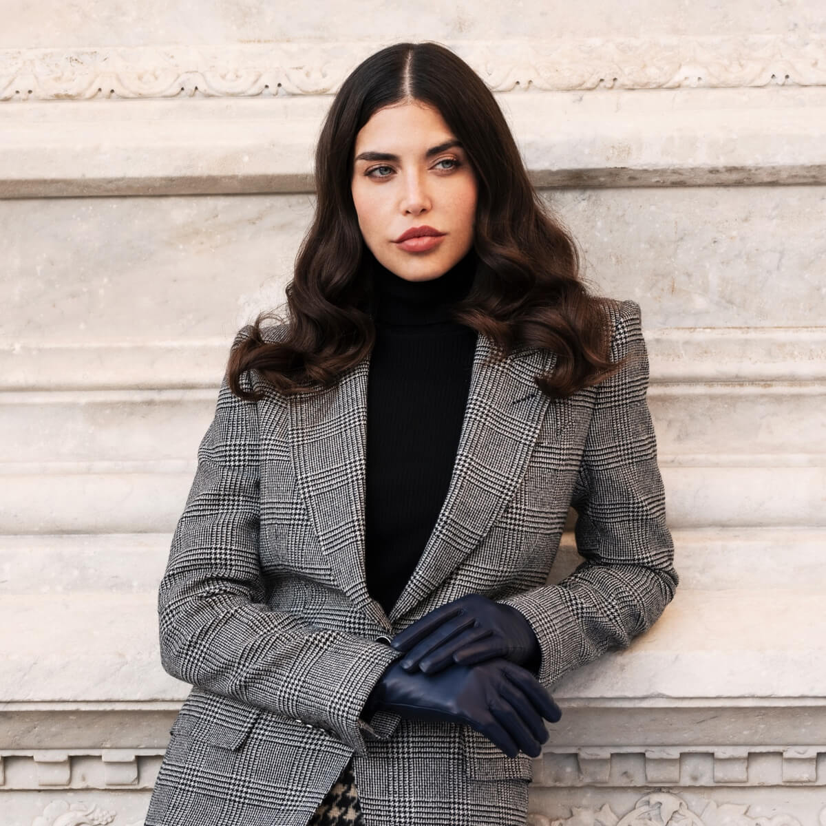 Woman wearing a gray checkered blazer and black gloves against a light stone wall.