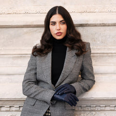 Woman wearing a gray checkered blazer and black gloves against a light stone wall.