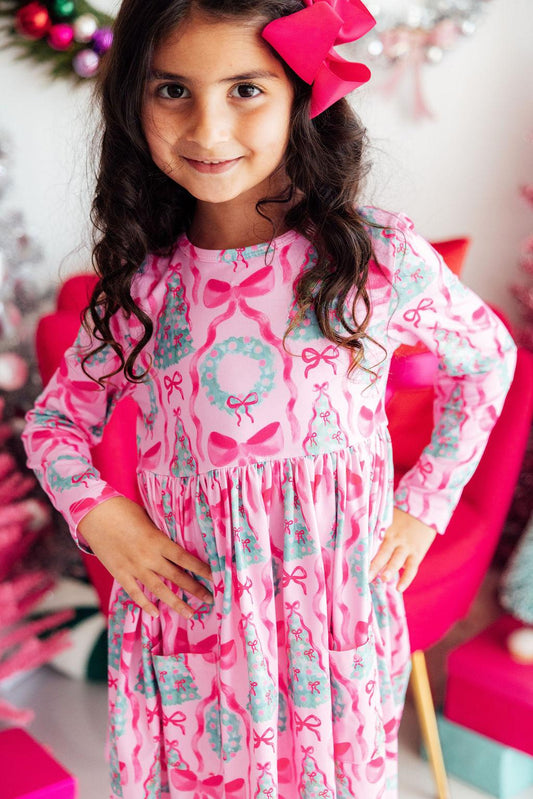 A young girl with long dark curly hair wearing a pink long-sleeve dress with a holiday print of bows, wreaths, and Christmas trees, accessorized with a large pink bow in her hair.