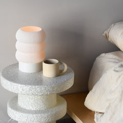 Small stone table with a textured lamp and mug next to a bed.