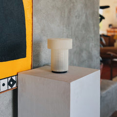 White ribbed vase on a white pedestal against a textured wall with abstract art.