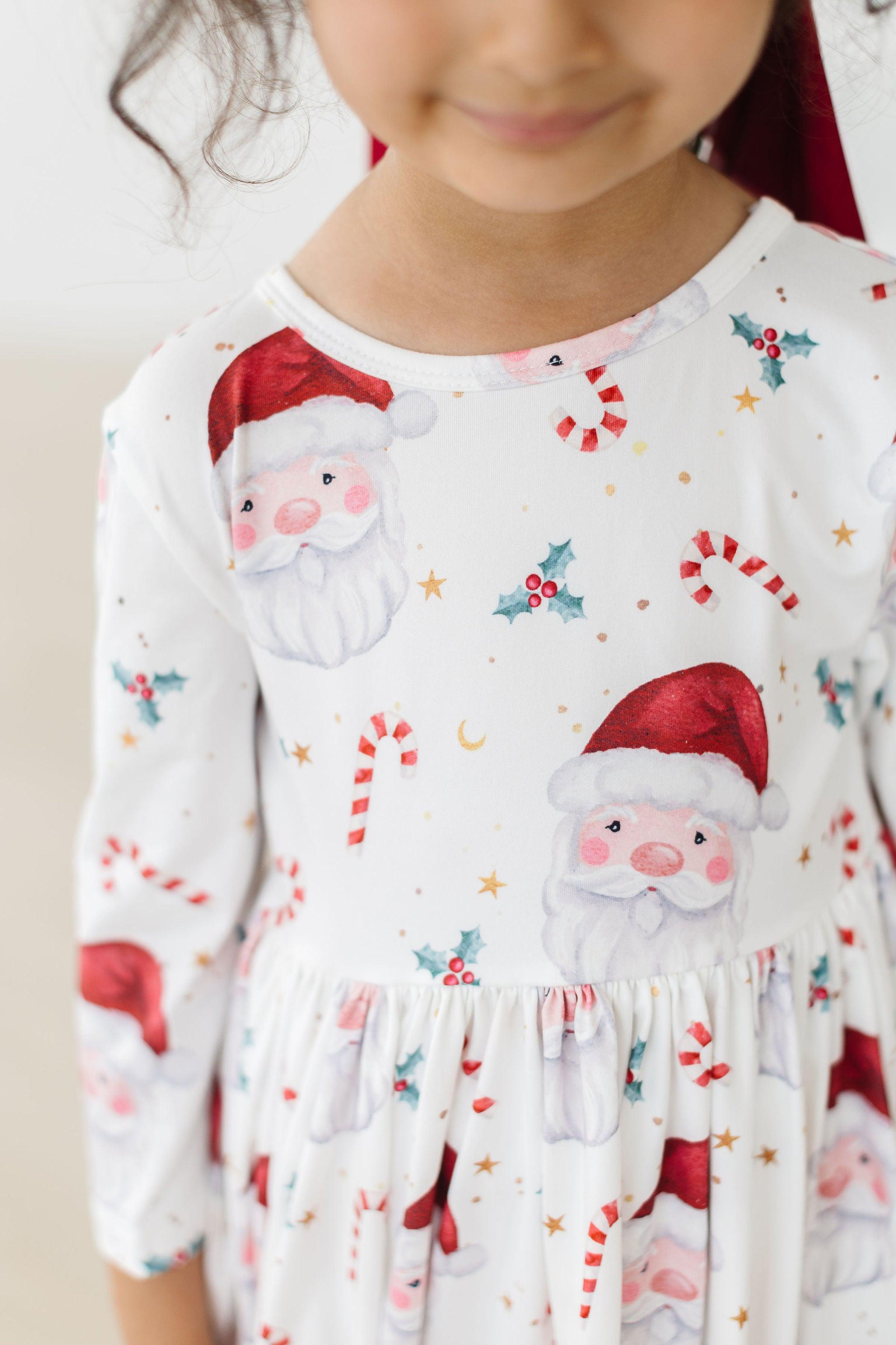 A close-up of a child wearing a white long-sleeve dress with a festive Christmas print featuring Santa Claus faces, candy canes, holly, and gold stars.
