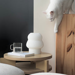 White cat on a wooden shelf with a round wooden table featuring a vase and glass.