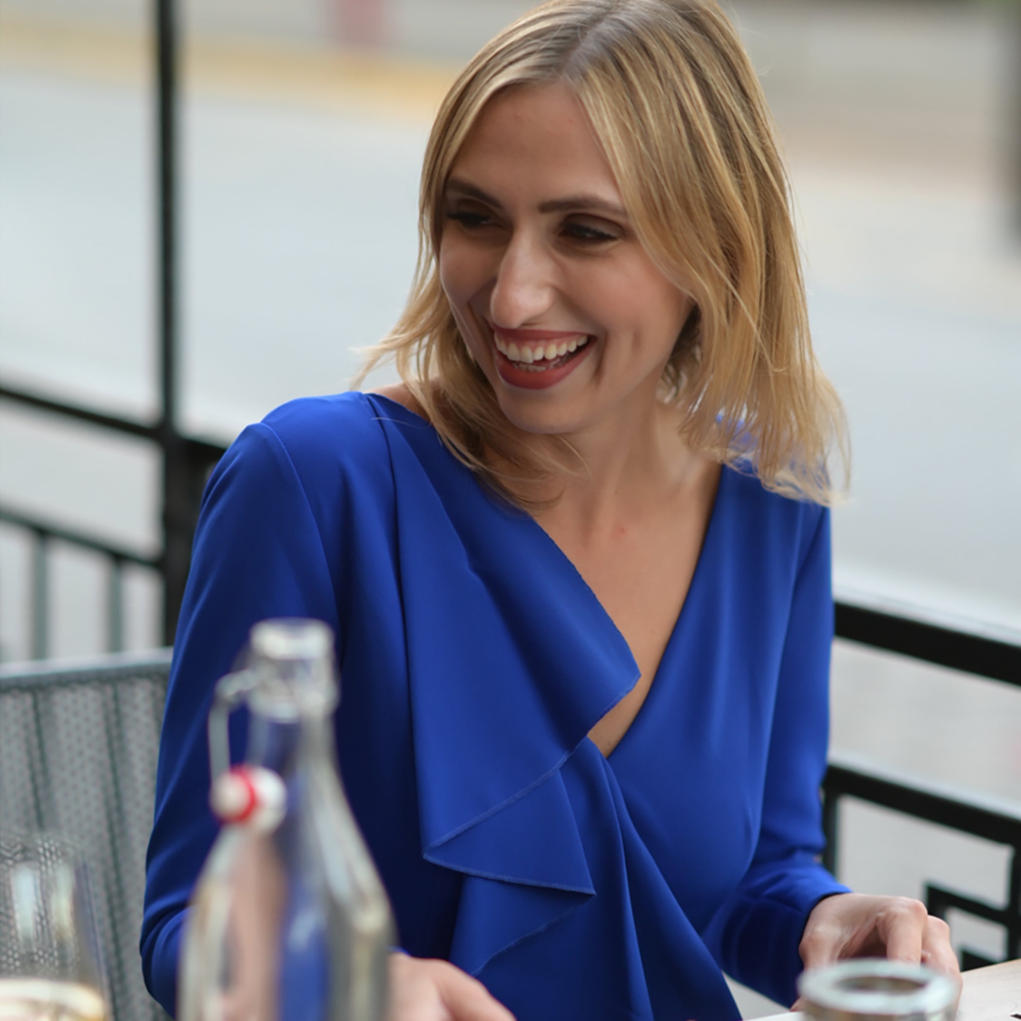 Woman in a blue dress smiling at a table with bottles and glasses.