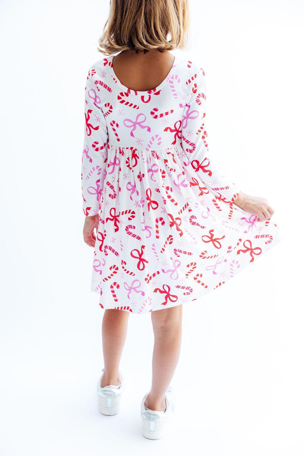 A child wearing a white long-sleeve dress with a red and pink candy cane and bow pattern, shown from the back against a white studio background.
