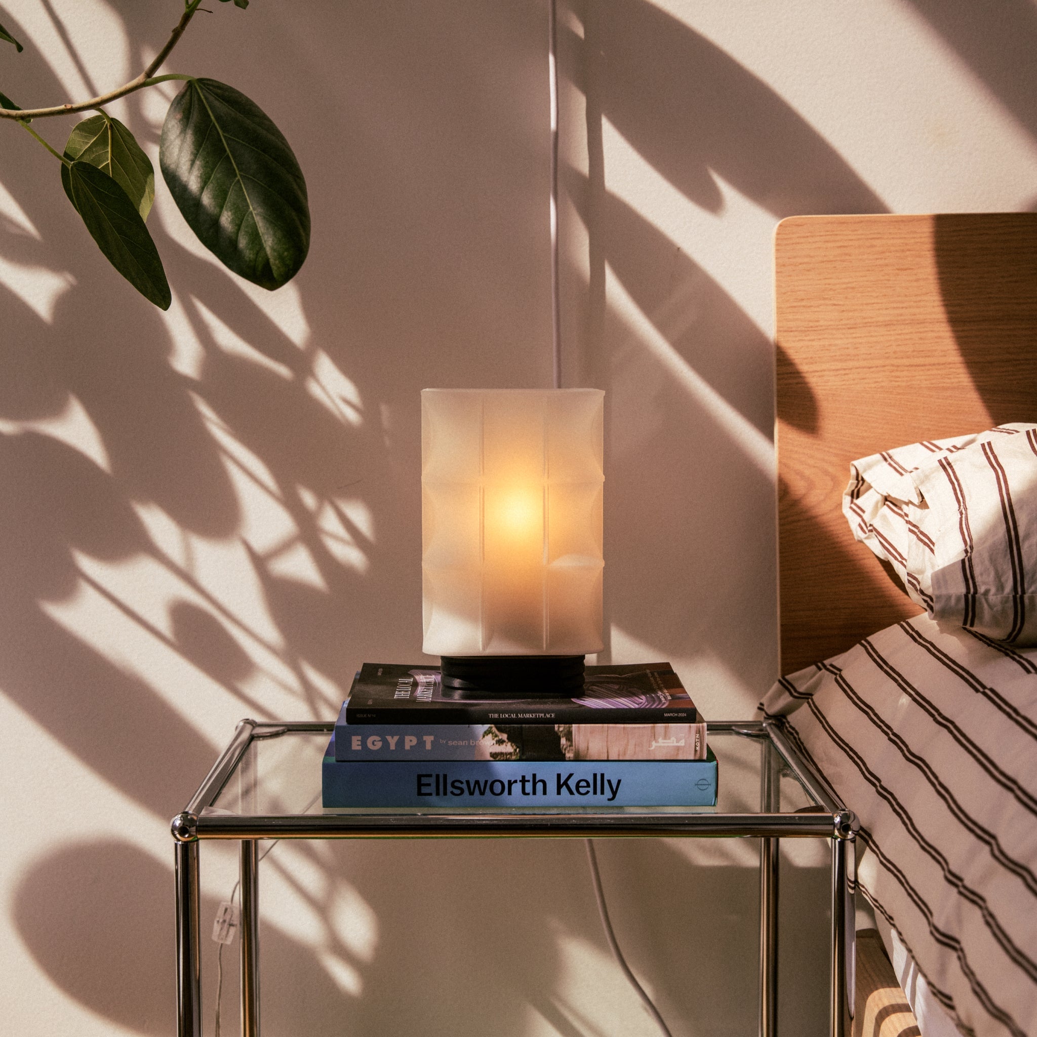 Glass nightstand with books and a lamp next to a bed with striped bedding.