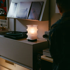 Person standing in a room with a lamp on a surface and framed pictures in the background