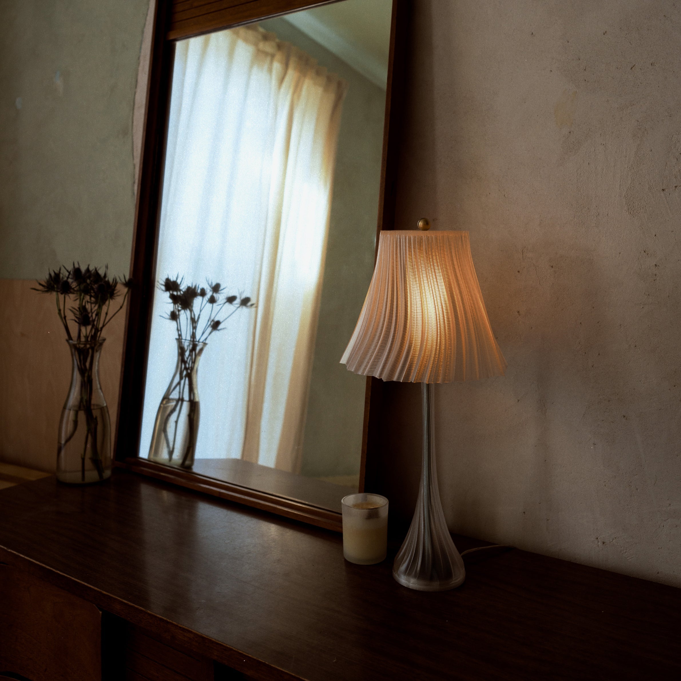 Lamp on a table next to a mirror reflecting a window with curtains.