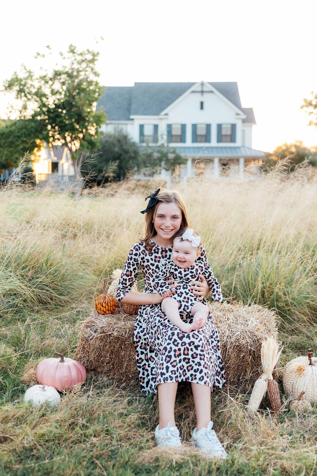 Woman and child sitting on a hay bale in a field with pumpkins and a house in the background