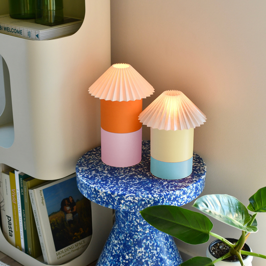 Two colorful table lamps on a blue speckled pedestal with a bookshelf and plant in the background.