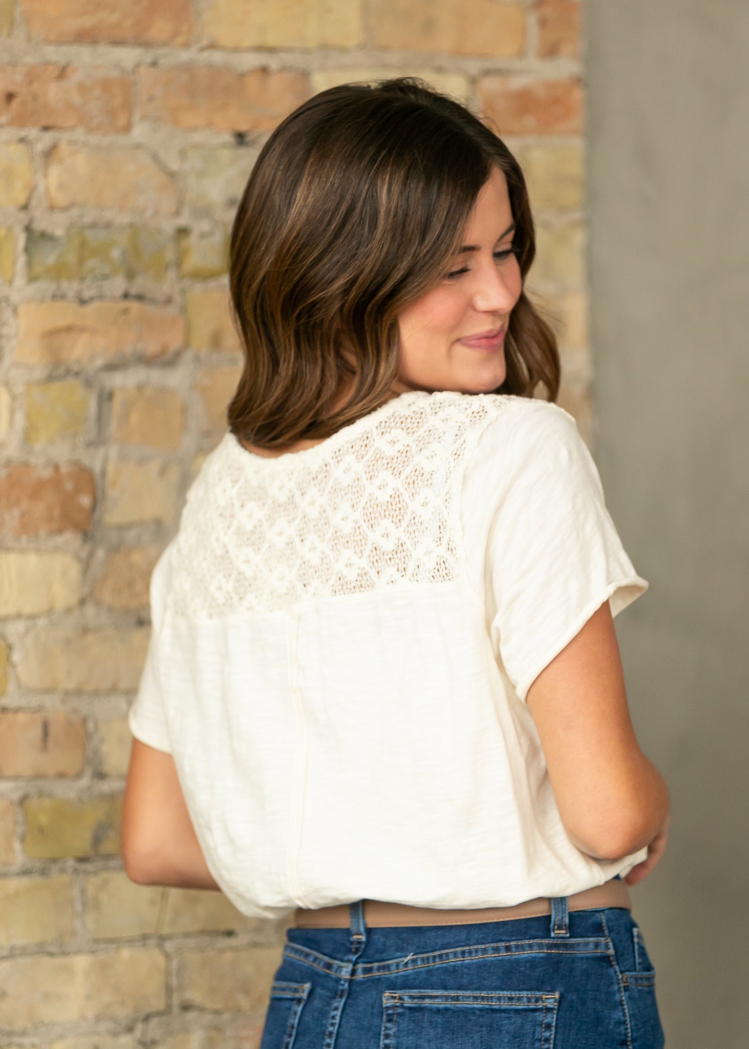 Woman wearing a white lace top and blue jeans against a brick wall.