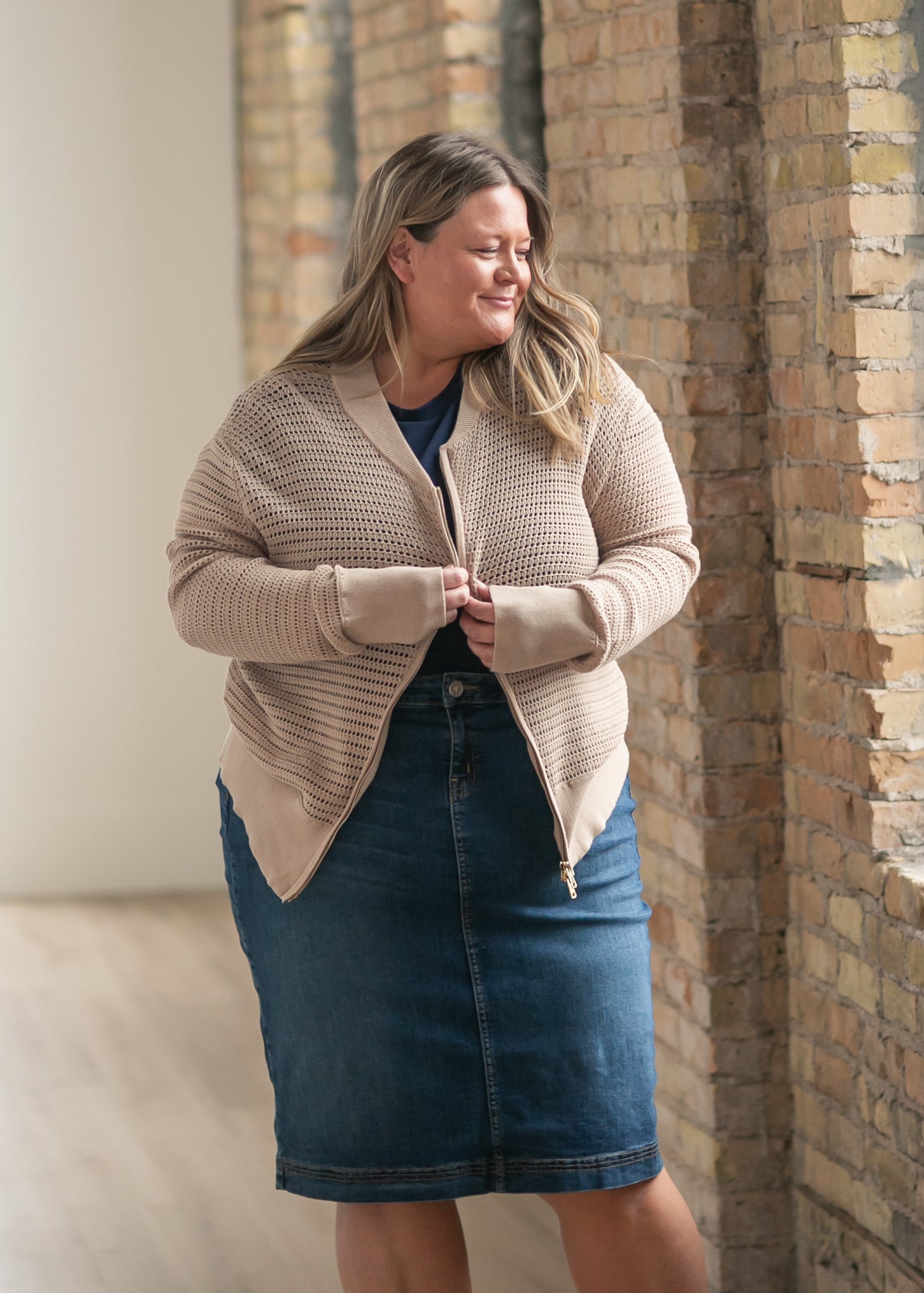 Woman wearing a beige cardigan and blue skirt standing against a brick wall.