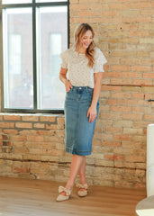 Woman in a white blouse and blue denim skirt standing against a brick wall.