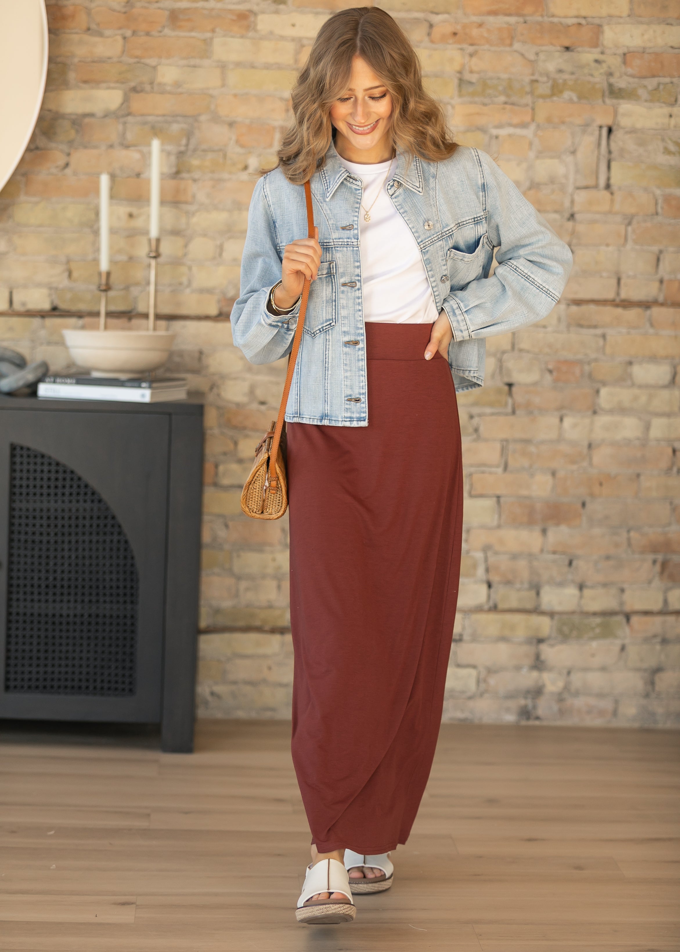Woman in a denim jacket and brown skirt standing in a room with a stone wall.