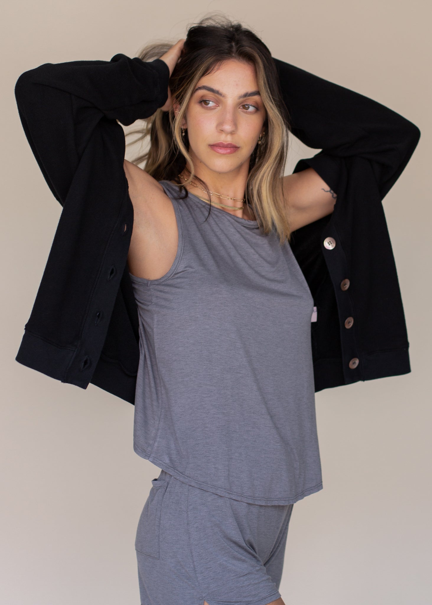 Woman wearing a gray sleeveless top and pants with a black jacket over her shoulder against a beige background