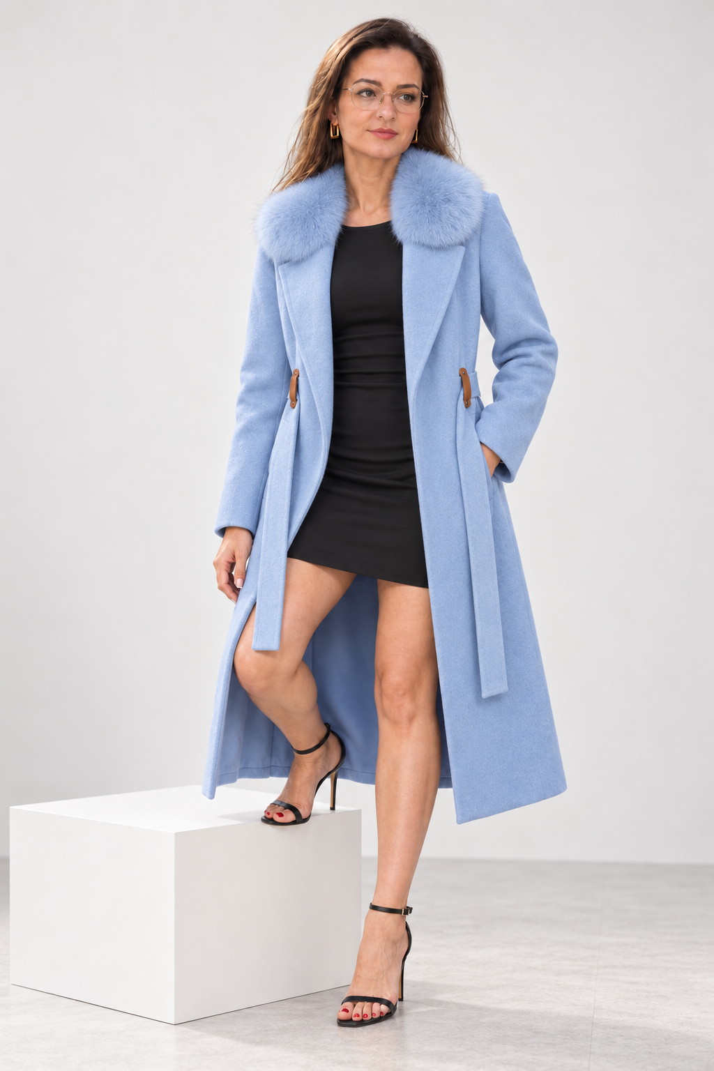 A woman models a light blue mid-length coat with a fluffy fur-trimmed collar, worn over a black mini dress and black high-heeled sandals, standing on a white display block against a light gray studio background.