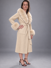 A woman models a beige wool coat with a thick cream fur collar and matching fur-trimmed cuffs, paired with black high-heeled sandals against a neutral gray studio background.