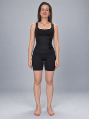 A woman stands barefoot against a light gray studio background wearing a black waist trainer vest with matching black fitted shorts.