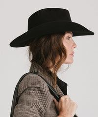Woman wearing a black wide-brimmed hat and holding a bag against a plain background