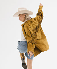 Woman wearing a mustard yellow fringed jacket and denim shorts on a white background