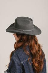 Person wearing a dark gray fedora hat and denim jacket on a plain background