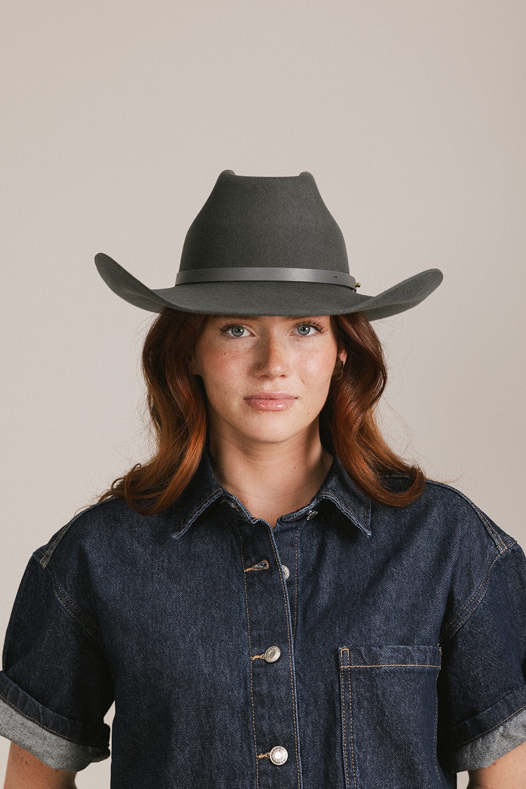 Woman wearing a dark gray fedora hat and denim shirt against a plain background
