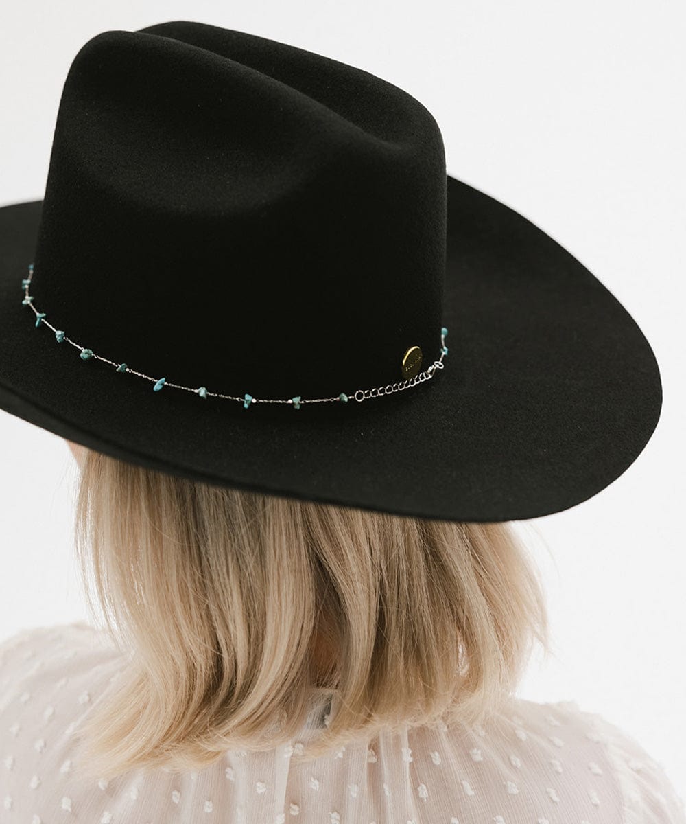 Black fedora hat with decorative band worn by a person with blonde hair on a white background