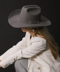 Person wearing a gray cowboy hat and light-colored coat against a black background