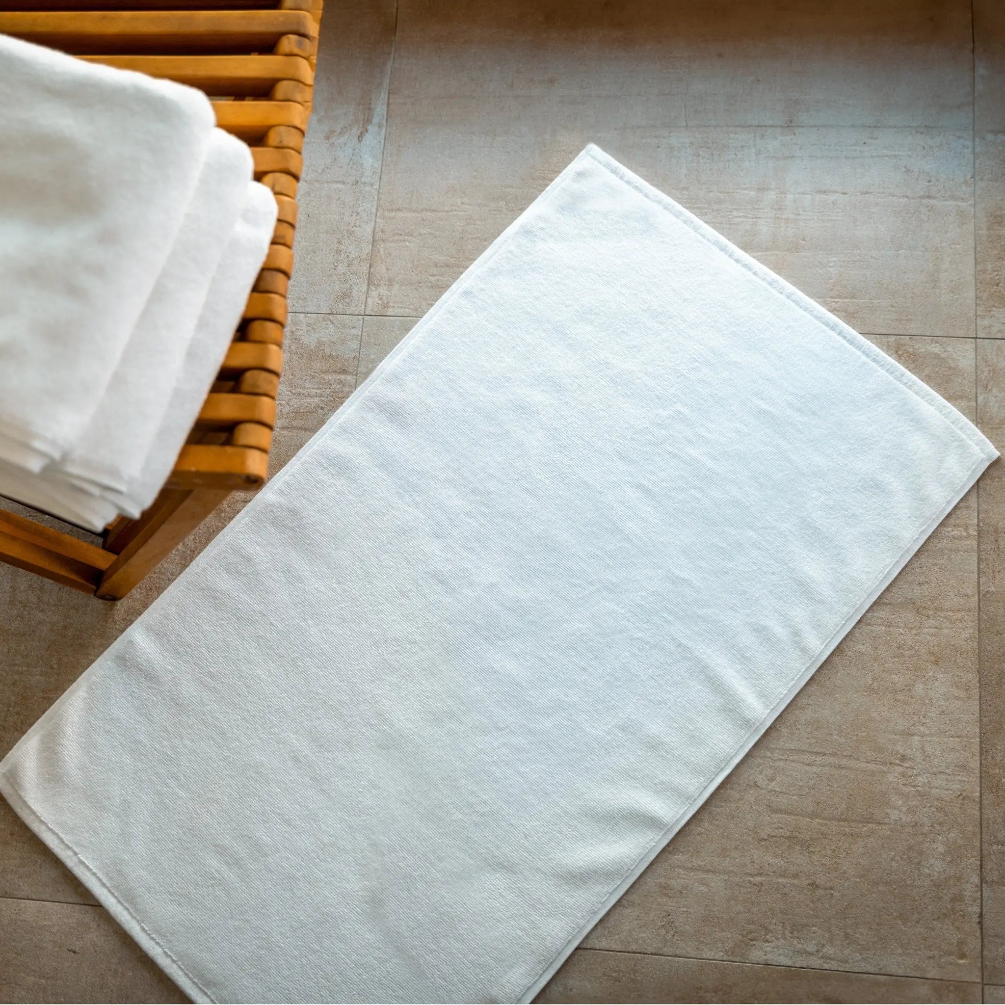 White bath mat on a tiled bathroom floor with a wooden basket containing additional white towels.