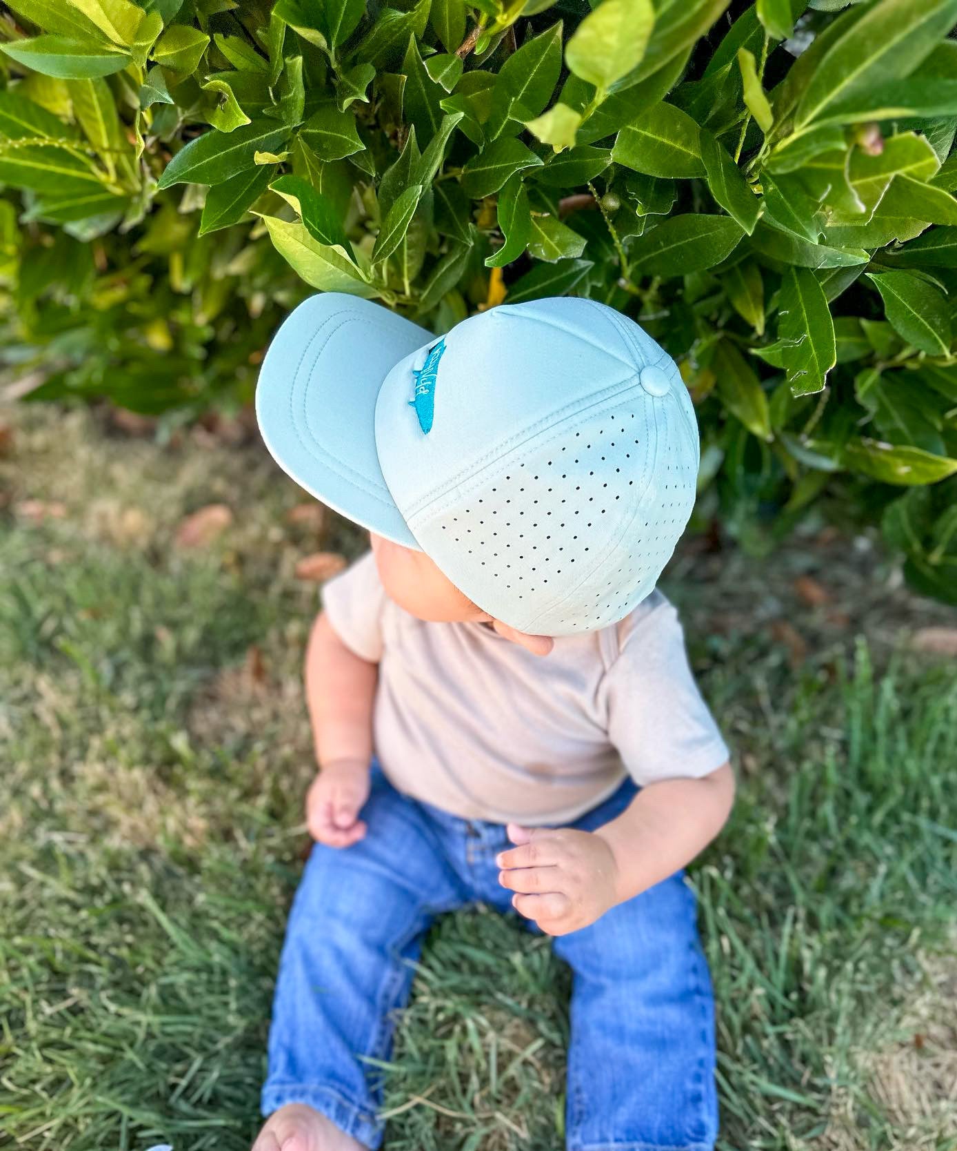 Child wearing two light blue caps outdoors on grass with green bushes in the background