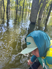 Person wearing a blue cap and life jacket in a flooded area with trees reflected in the water.
