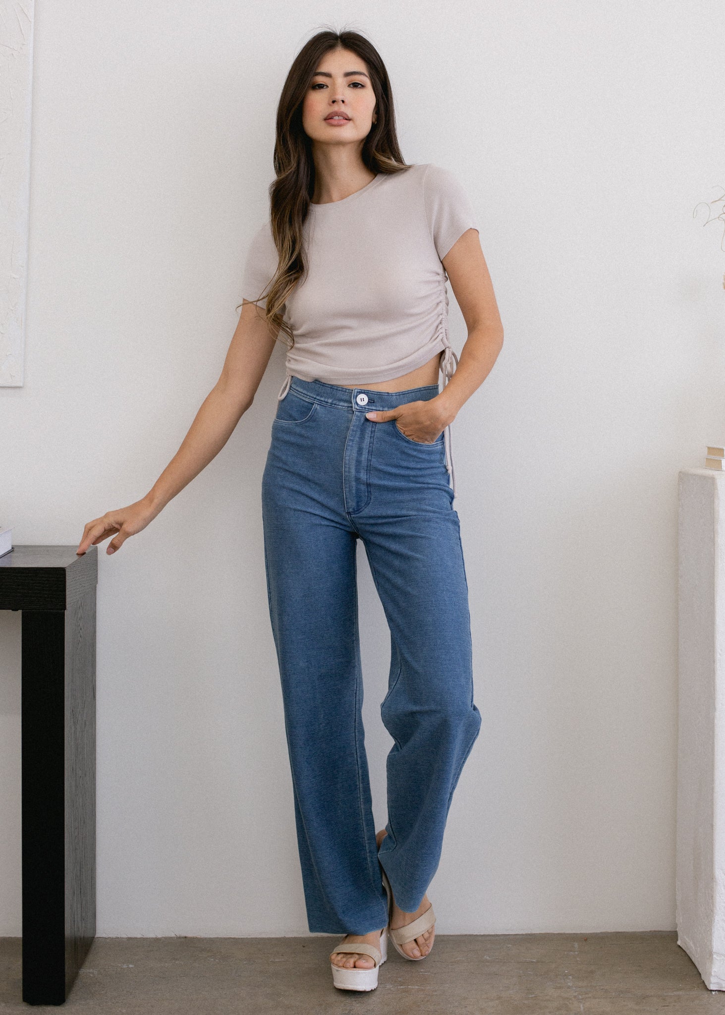 Woman wearing a beige top and blue jeans standing against a white wall.