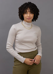 Woman wearing a beige turtleneck and green pants against a gray background