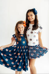 Two young girls modeling matching sports-themed apparel against a plain white studio background. The girl on the left wears a navy blue sleeveless dress with a dog face print, and the girl on the right wears a white short-sleeve top with dog prints, a navy gingham skirt, and a navy hair bow.