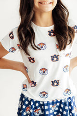 A woman wearing a white short-sleeve t-shirt with an all-over Auburn Tigers logo print, paired with a blue and white gingham skirt. The Auburn 'AU' logo and tiger mascot graphics are printed across the shirt.