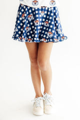 A person wearing a blue and white gingham skater skirt with a repeating pattern of cartoon tiger faces, paired with white sneakers, shown from the waist down on a white background.